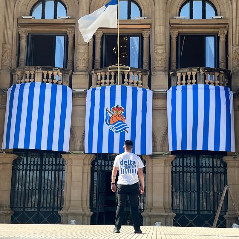 Balcones de un edificio histórico de Donostia con lonas blanquiazules impresas por Delta y la camiseta de Delta en primer plano