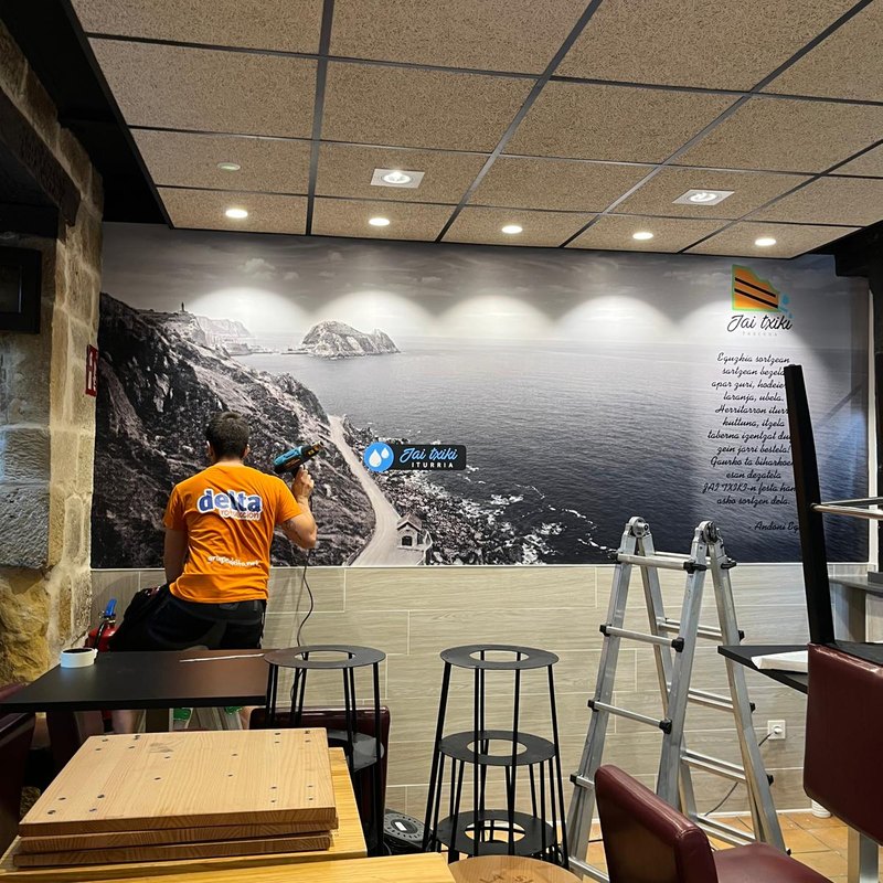 Instalador de Delta colocando un mural vinilado con un paisaje de costa en la pared de un bar