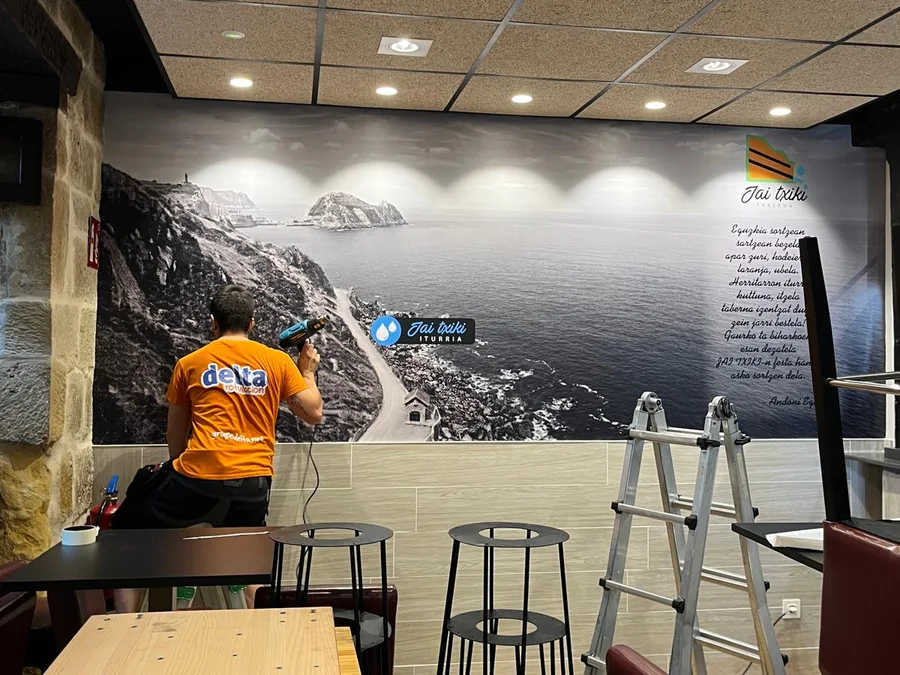 Instalador de Delta colocando un mural vinilado con un paisaje de costa en la pared de un bar